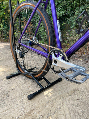 A close-up outdoor shot of a purple bicycle frame and rear wheel resting on a black stand, with a silver crankset and gray pedal visible against a gravel path and green foliage background.